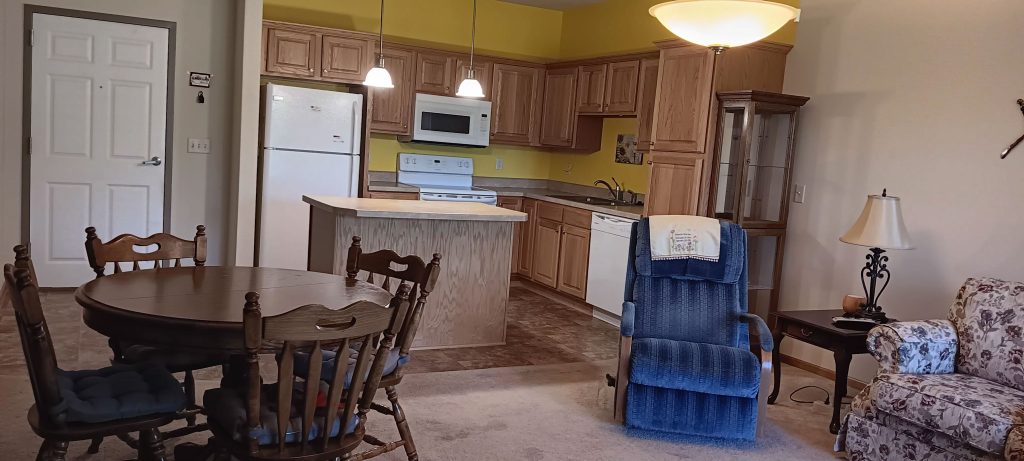 A cozy kitchen and dining area with wooden cabinets, a white refrigerator, stove, and microwave. A small dining table with chairs is near a blue recliner and a floral sofa. Soft lighting and beige carpet create a warm atmosphere.