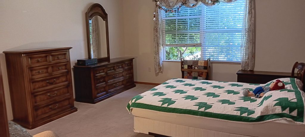 A bedroom with a bed covered in a green and white quilt, featuring a teddy bear and doll. There's a wooden dresser with a mirror, another dresser, a chair, and a large window with blinds and curtains.
