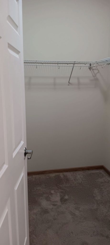 A walk-in closet with an open door. The closet features an empty white wire shelf along two walls and a brown carpeted floor. The walls are light-colored, and the closet is empty, suggesting ample storage space.