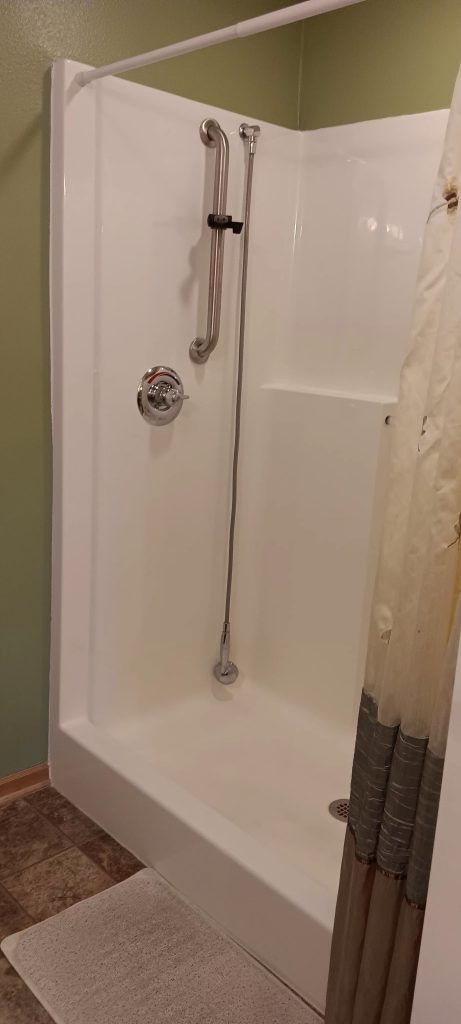 A shower area with a white stall, equipped with a vertical grab bar and a handheld showerhead. The curtain is partially drawn, revealing a green wall and a section of tiled floor with a white bath mat.