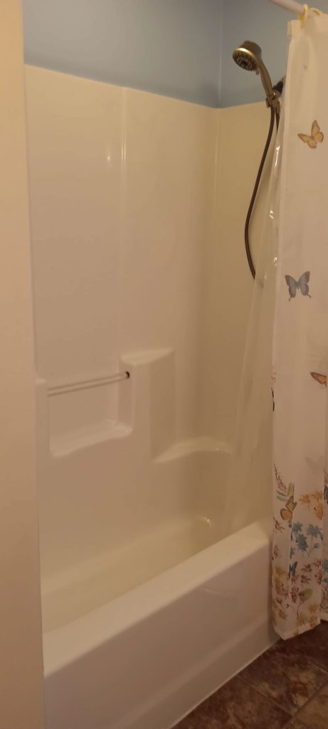 A bathroom with a white bathtub and shower combination. It features a mounted showerhead with a dark hose. The shower curtain is decorated with colorful butterflies. The tub has built-in shelves and a small handrail. The floor is brown patterned.
