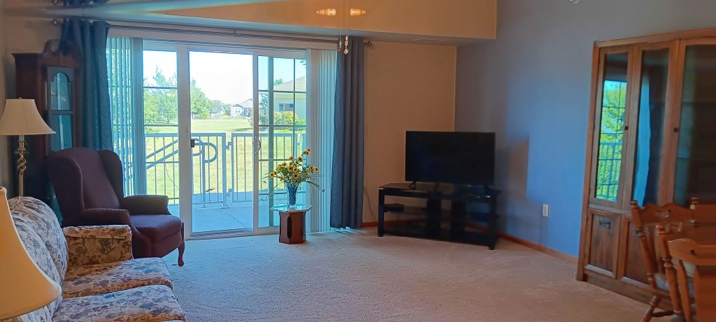 A living room with a large window leading to a patio. There is a sofa, an armchair, a TV on a stand, and a wooden cabinet. A small table holds a vase with yellow flowers. The room has beige carpeting and light walls.