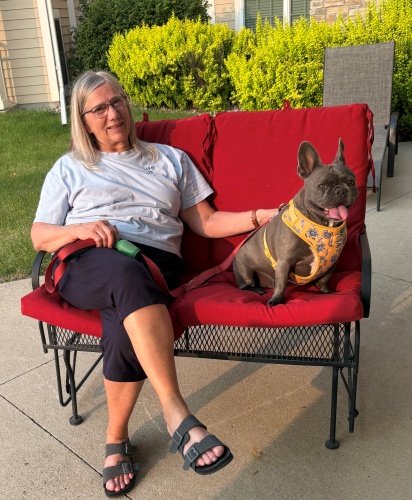 Anna with gray hair sits on a red cushioned outdoor bench, smiling and holding a leash attached to a gray French Bulldog wearing a yellow harness. They are on a patio with green bushes in the background.